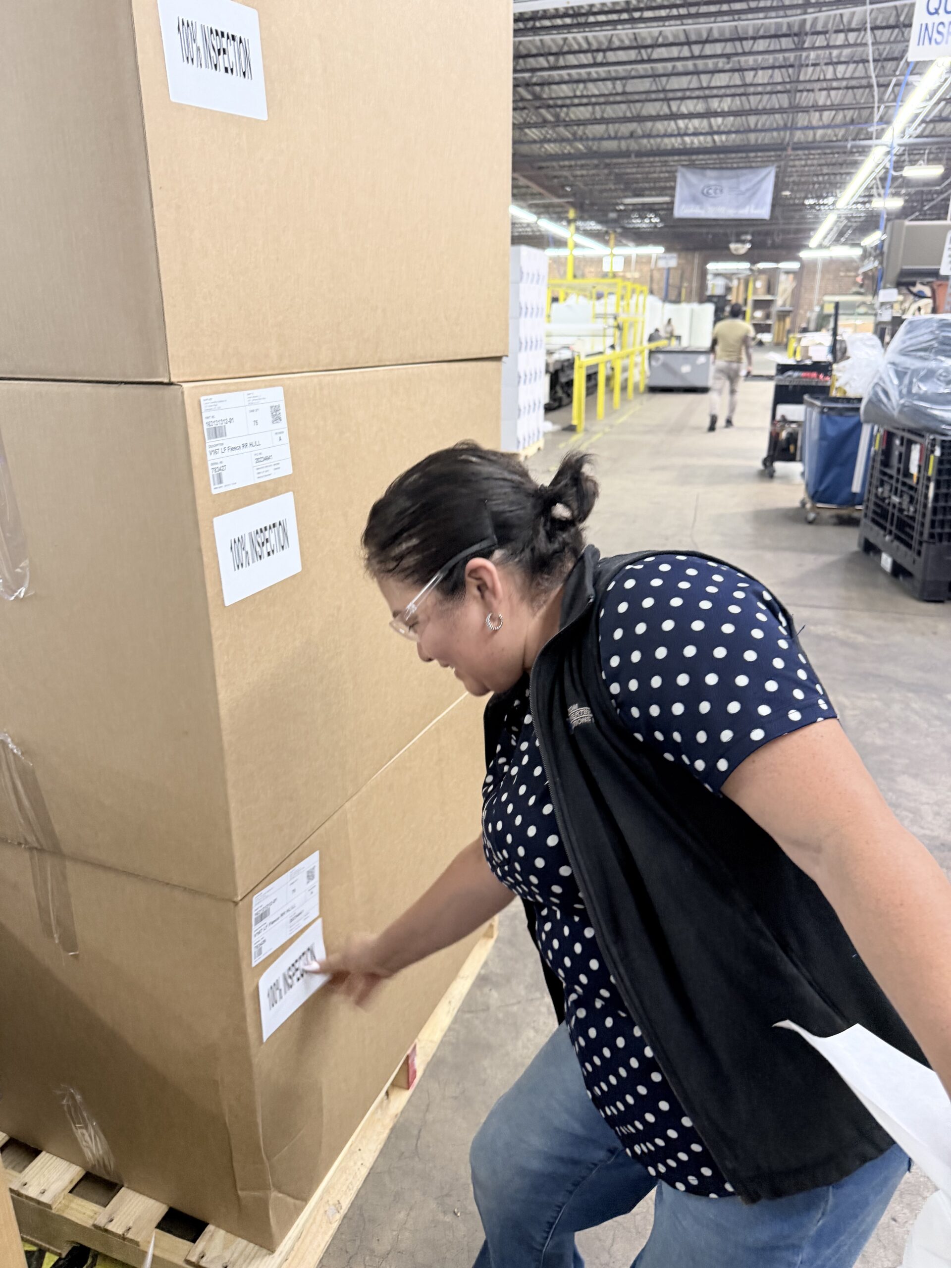 Woman inspecting a large cardboard box labeled "100% INSPECTION" in a warehouse setting, ensuring quality control on shipments.